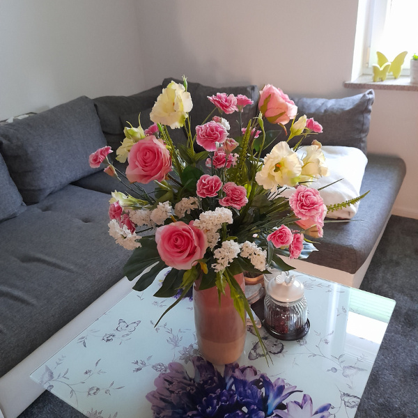 Wohnzimmer mit Kunstblumen aus rosa Rosen weiße Nelken und Gräser in Keramikvase auf Couchetisch neben Sofa